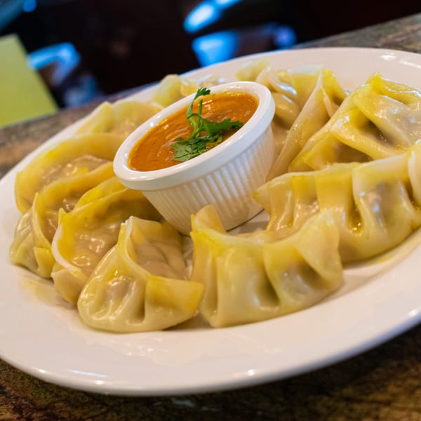 A plate of Guras Spice House momos, with a small bowl of orange dipping sauce garnished with cilantro.