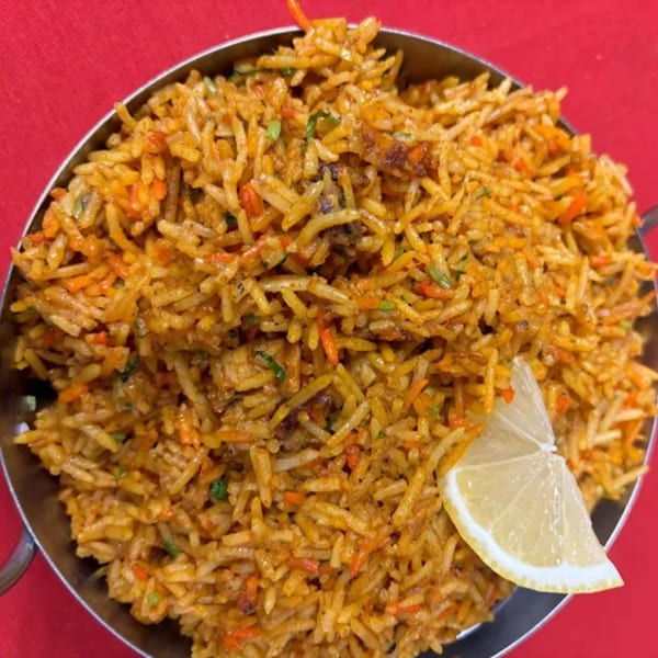 Indian biryani with rice, vegetables, and a lemon wedge in a metal bowl on a red tablecloth.